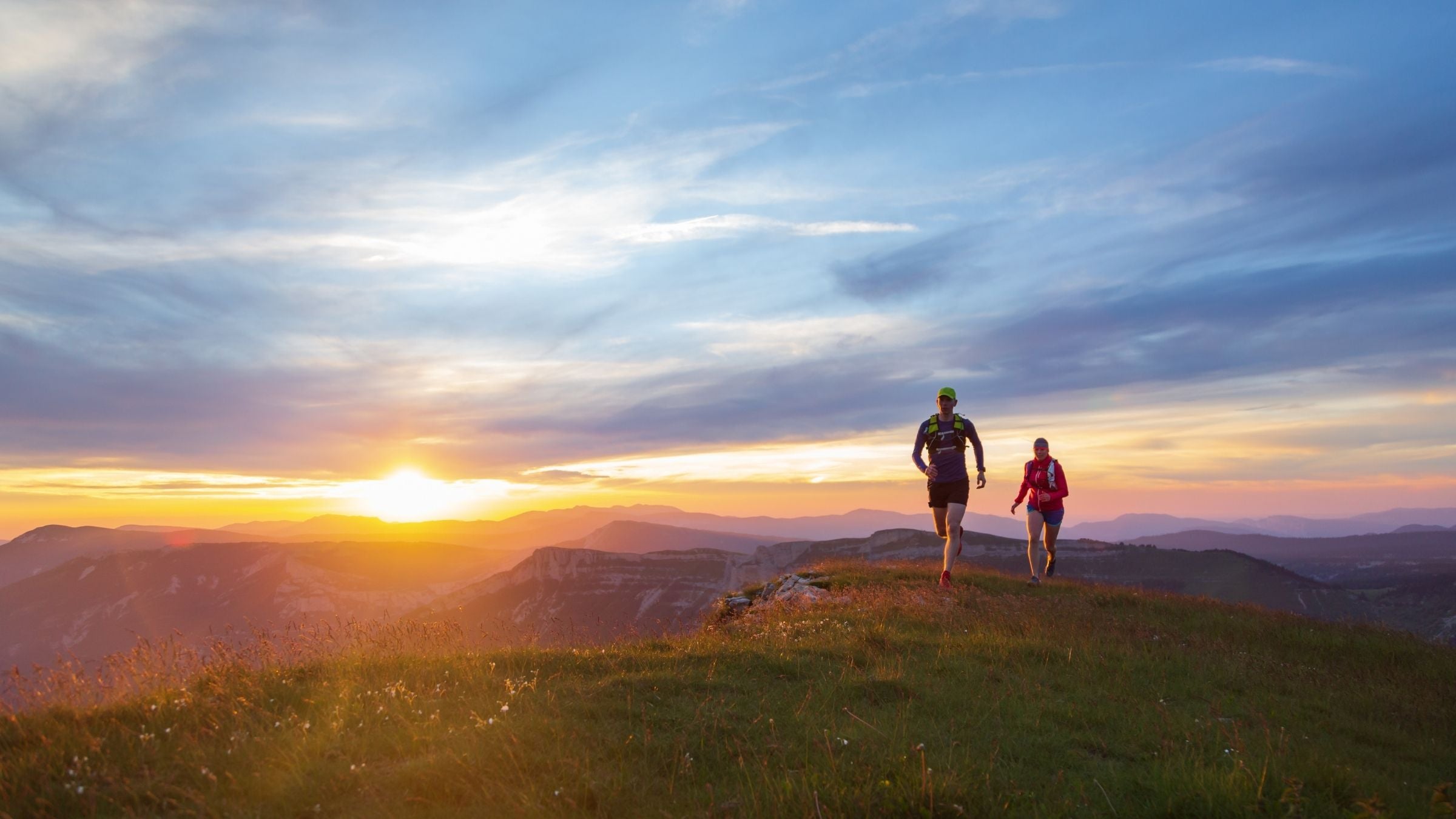 Two runners on a trail at a high elevation. The sun is rising in the background.