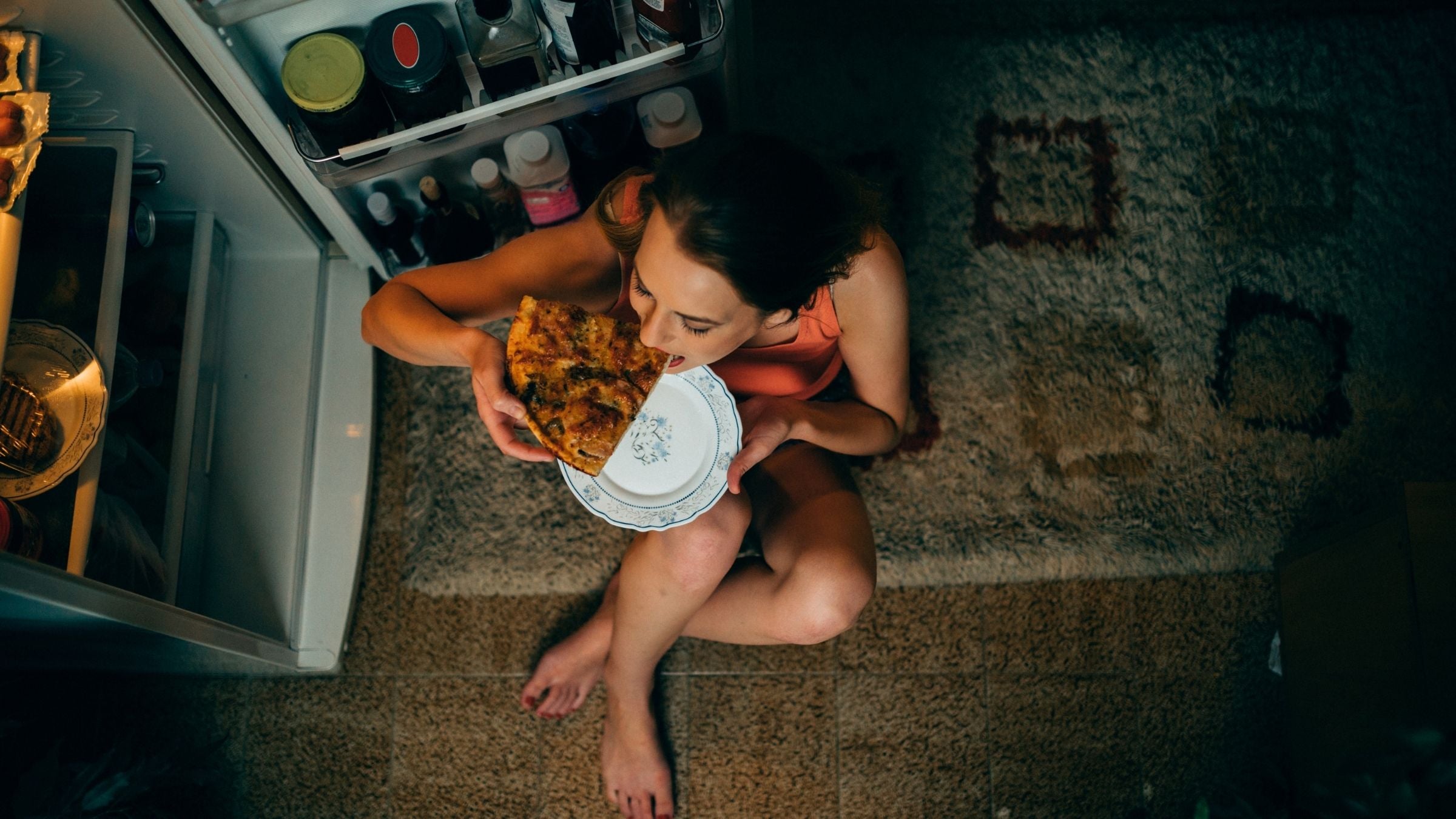 A woman sits in front of her open fridge, eating a slice of pizza.