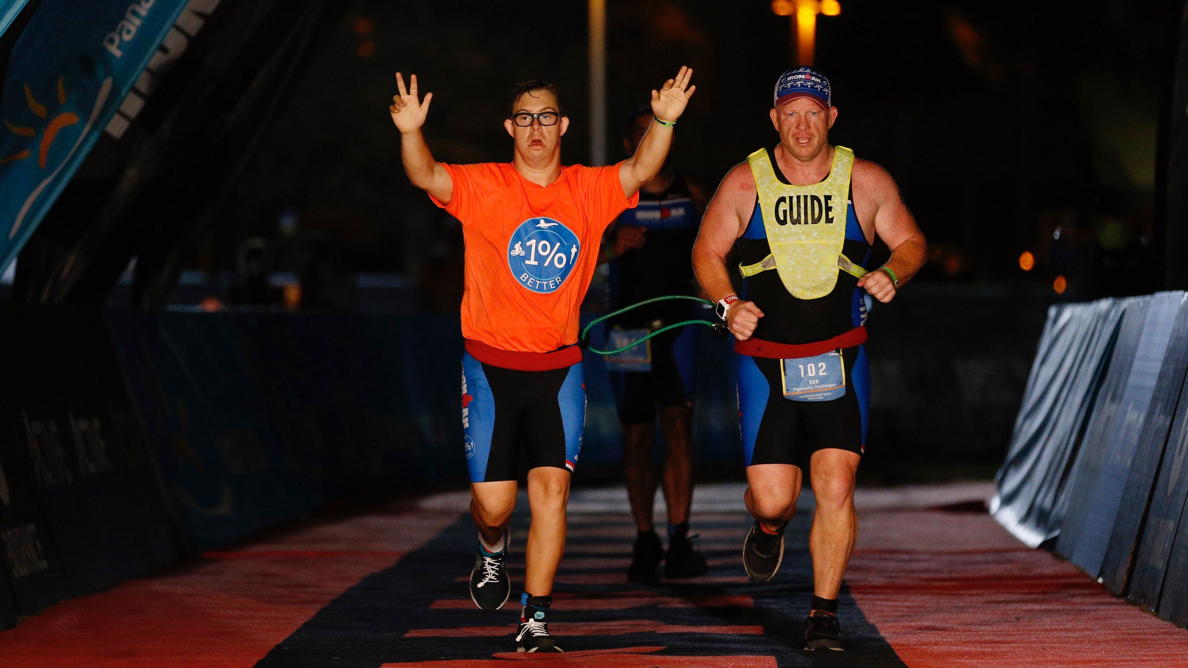 A young man with Down syndrome crosses the finish line of an Ironman triathlon with his arms in the air. He is tethered to an older man who serves as his guide.