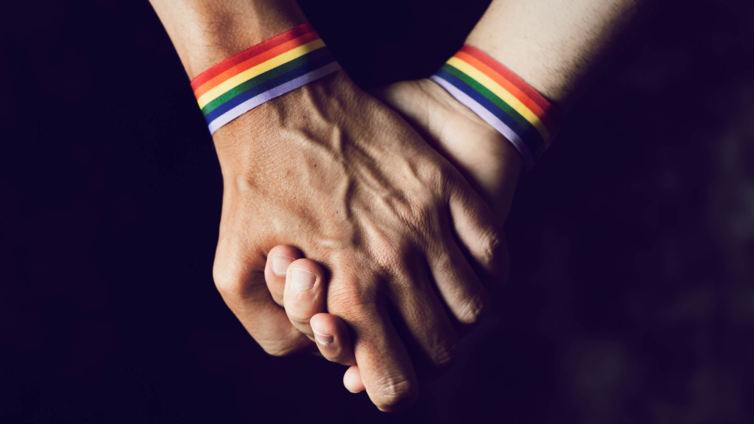 Closeup of two Caucasian men holding hands with a rainbow-patterned wristband on their wrists