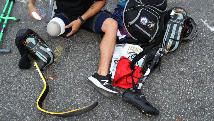 A man sits on the ground while preparing his amputated leg to receive a prosthetic running blade. He is surrounded by gear to race a triathlon.