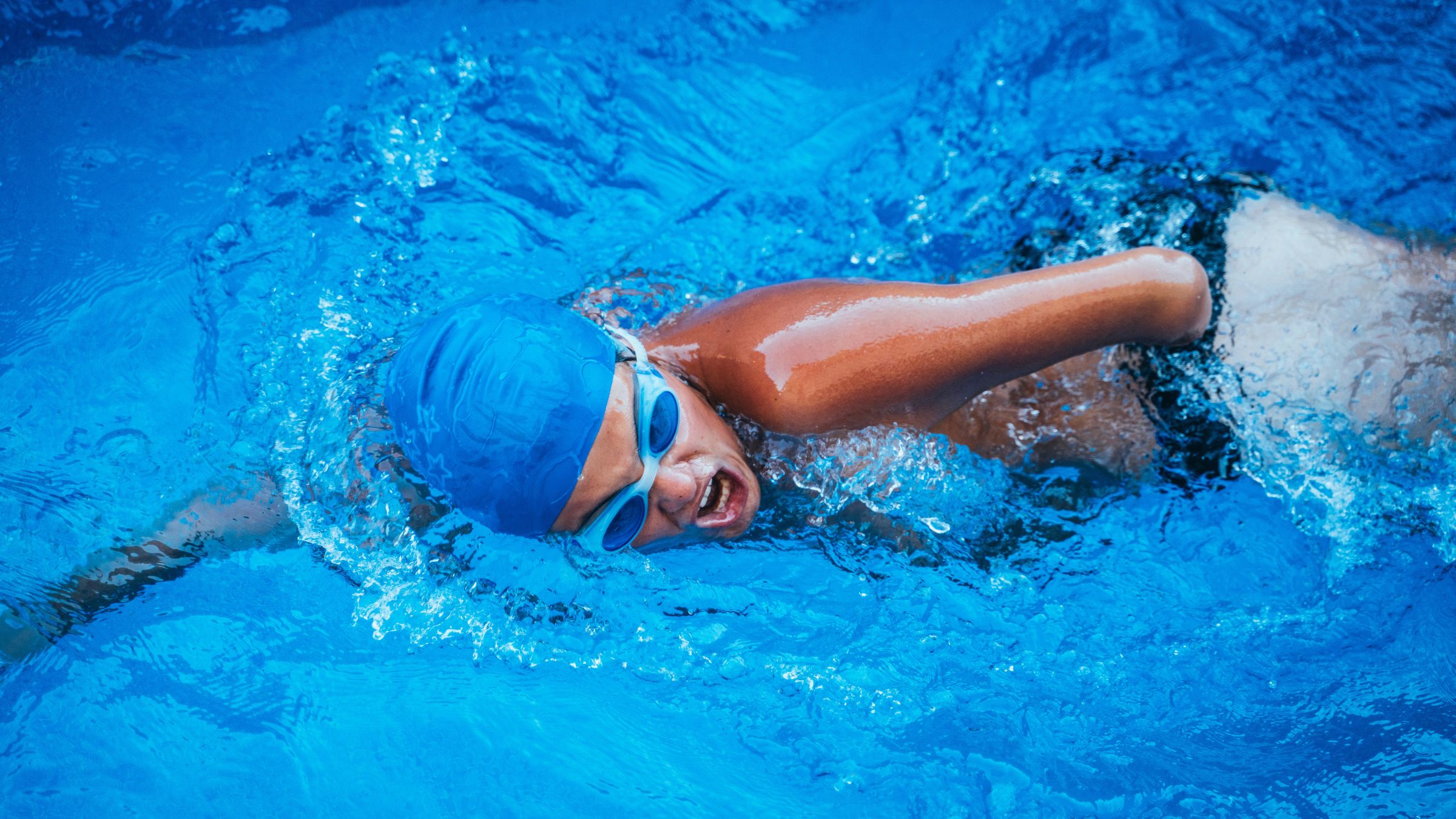 A male with a limb difference on her left arm rolls to her side to take a breath as she swims. He is wearing a black swimsuit, blue swim cap, and goggles.