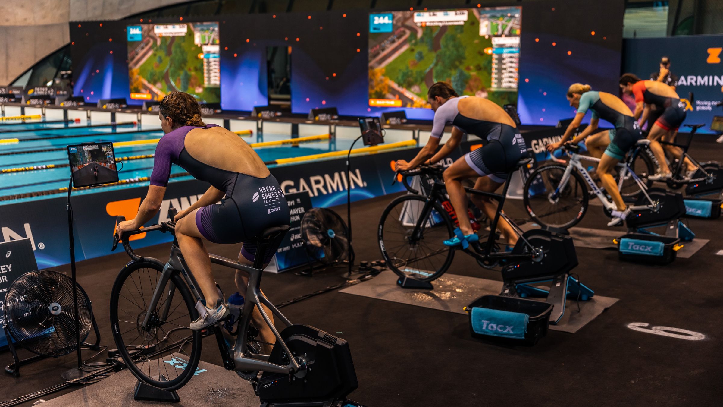 Athletes ride bikes in front of a video screen at Super League Arena Games, a video gamification triathlon