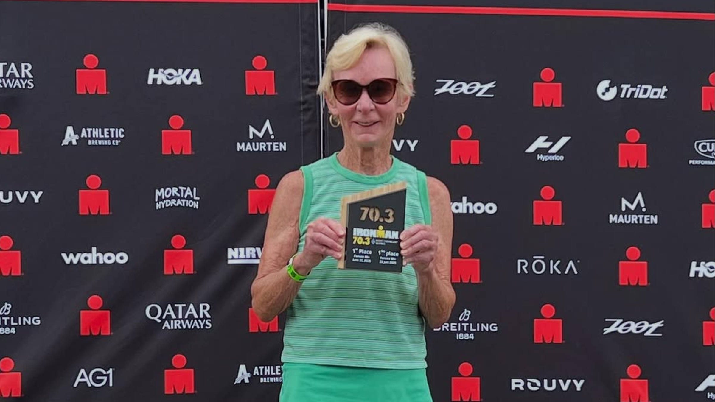 older woman stands in front of Ironman backdrop holding 70.3 award